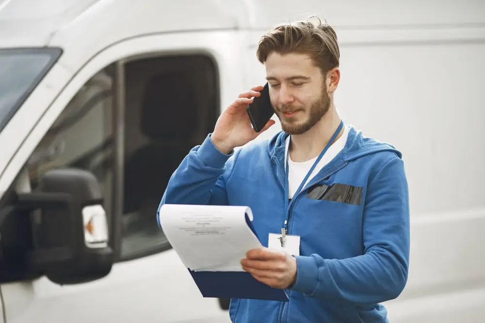 A tradesperson taking a phone call beside a work van, holding a job sheet