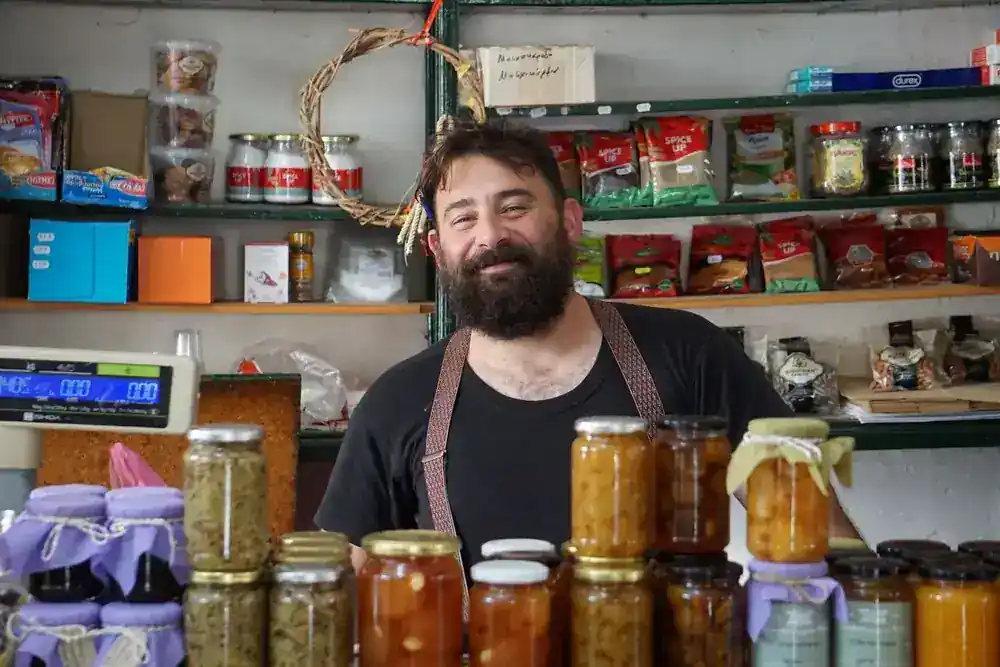 Smiling shopkeeper behind a counter of jarred preserves in a small local store
